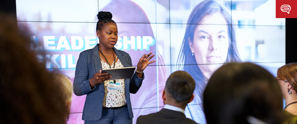 A woman stands holding a tablet and speaks to an audience; behind her, a large screen displays the words "LEADERSHIP SKILLS," a photo of another woman, and an executive summary slide.