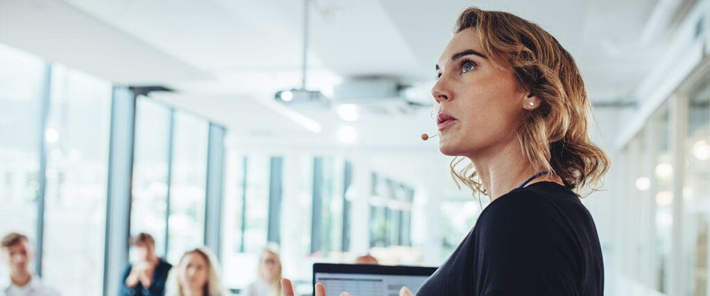 A woman wearing a headset microphone presents a healthcare pre-seed pitch deck to an attentive audience in a modern, bright conference room with large windows. People are seated and listening closely in the background.