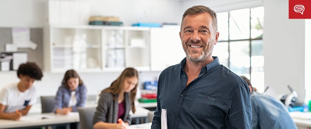 A smiling male teacher stands in a bright classroom holding papers, while four students work at desks in the background—perfect for illustrating focus and leadership in a healthcare pre-seed pitch deck. Shelves and a large window are visible behind him.