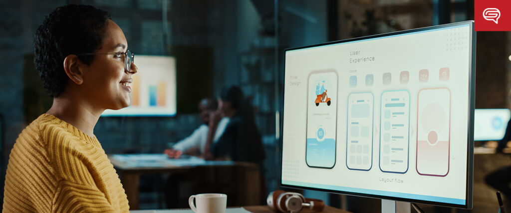 A person wearing glasses and a yellow sweater smiles while reviewing a healthcare pre-seed pitch deck on a monitor displaying user experience designs for a mobile app. A desk with headphones is in front of them, with two people blurred in the background.