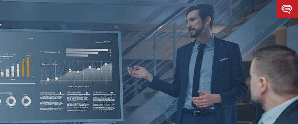 A man in a suit and tie stands next to a computer screen displaying pitch deck slides.