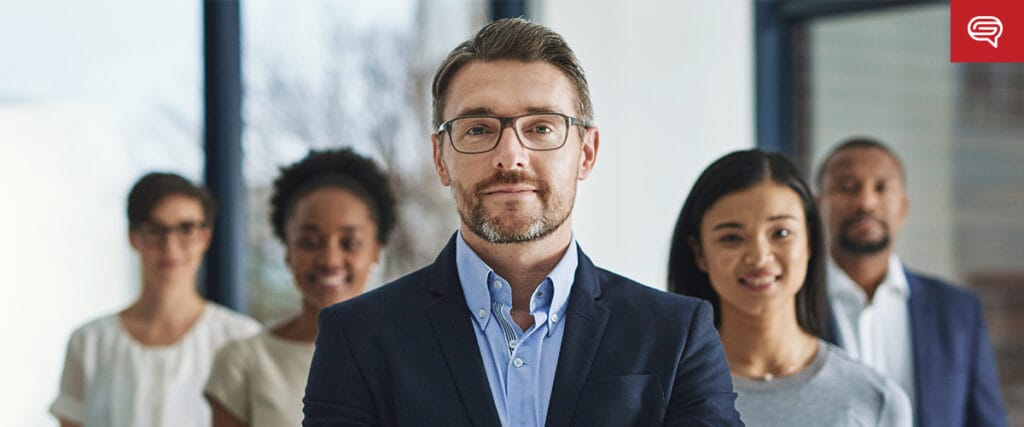 A confident man in a suit stands in front of a diverse group of four people, all smiling, during a brightly lit office business intelligence presentation.