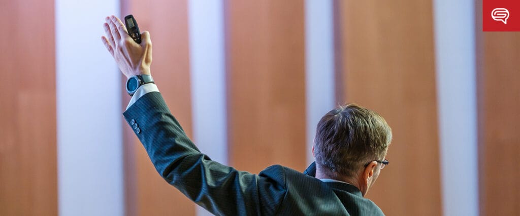 A man in a suit raises his left arm while presenting a medical slide, seen from behind. He holds a remote in his hand, and his audience is not visible. Vertical wooden panels are in the background.