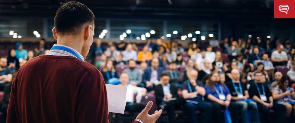A man holding papers presents a healthcare pre-seed pitch deck to a large seated audience in an auditorium, with people attentively listening and blurred stage lights in the background.