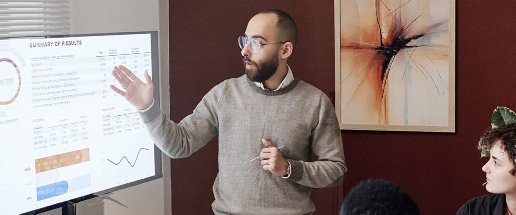 A man with glasses presents a healthcare pre-seed pitch deck, displaying charts and graphs on a large screen to a small group in a meeting room. Modern abstract art hangs on the wall behind him.