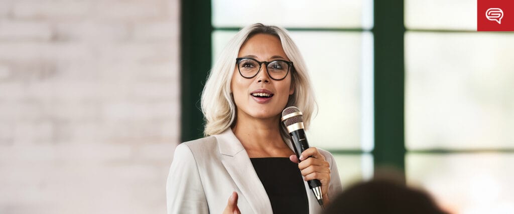 A woman with blonde hair and glasses, dressed in a white blazer and black top, confidently presents our value slide into a microphone in a bright room with large windows.