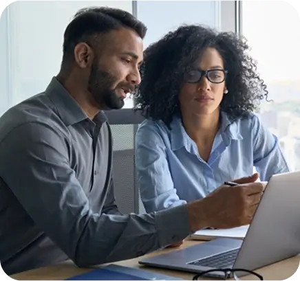 Two people sit at a desk in an office, looking at a laptop screen discussing an AI claims platform for life insurance. One person points at the laptop while explaining, and the other listens attentively. Both appear focused and engaged.